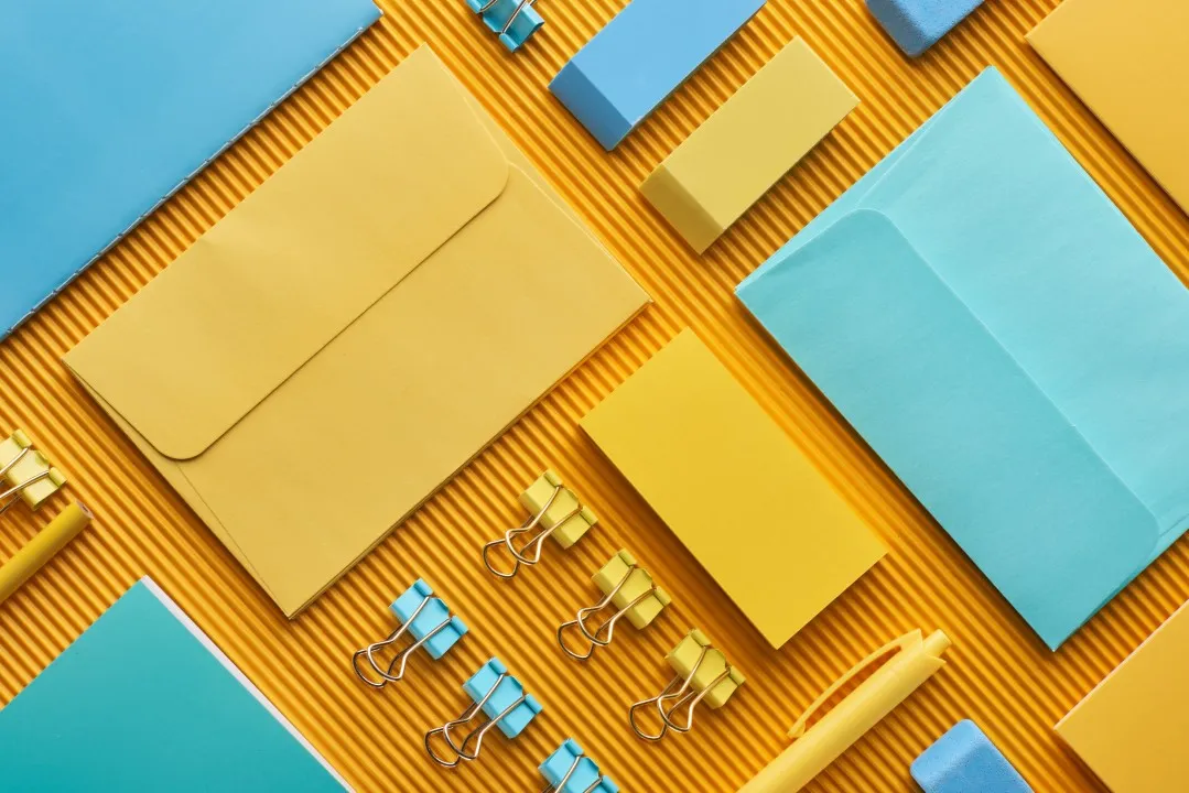 Set de papelería corporativa en amarillo y cian con clips, sobres y bolígrafo sobre fondo corrugado.